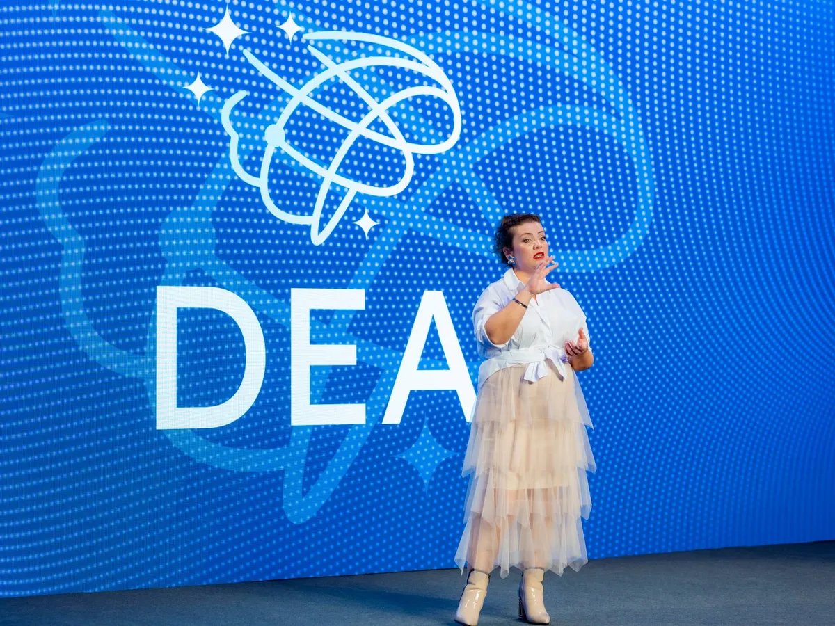 Irene Hernández hablando en un escenario delante de una gran pantalla azul con el logo de DEA